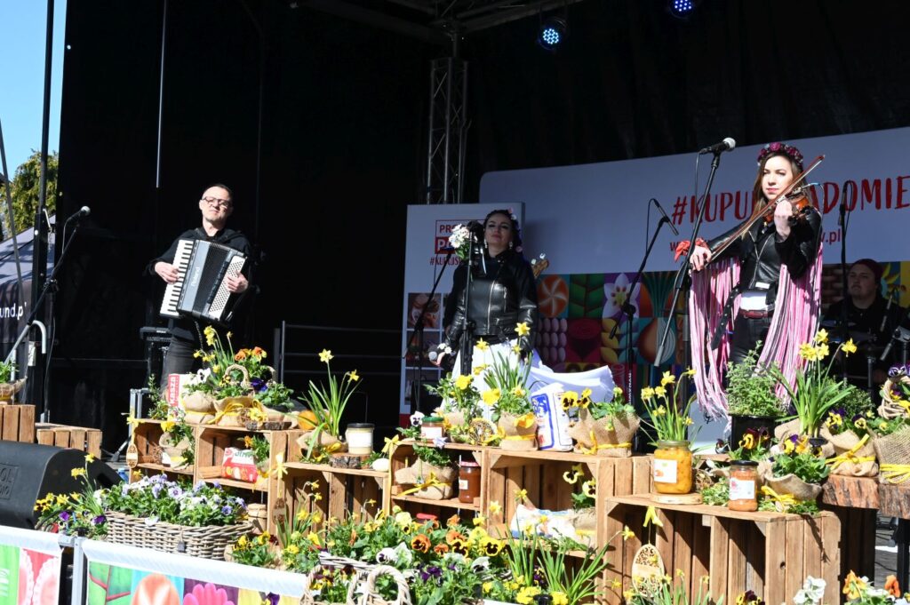 Muzycy na scenie podczas występu plenerowego – akordeonista oraz wokalistka i skrzypaczka, a przed sceną kolorowe stoiska z roślinami i rękodziełem (kliknięcie powiększy zdjęcie).