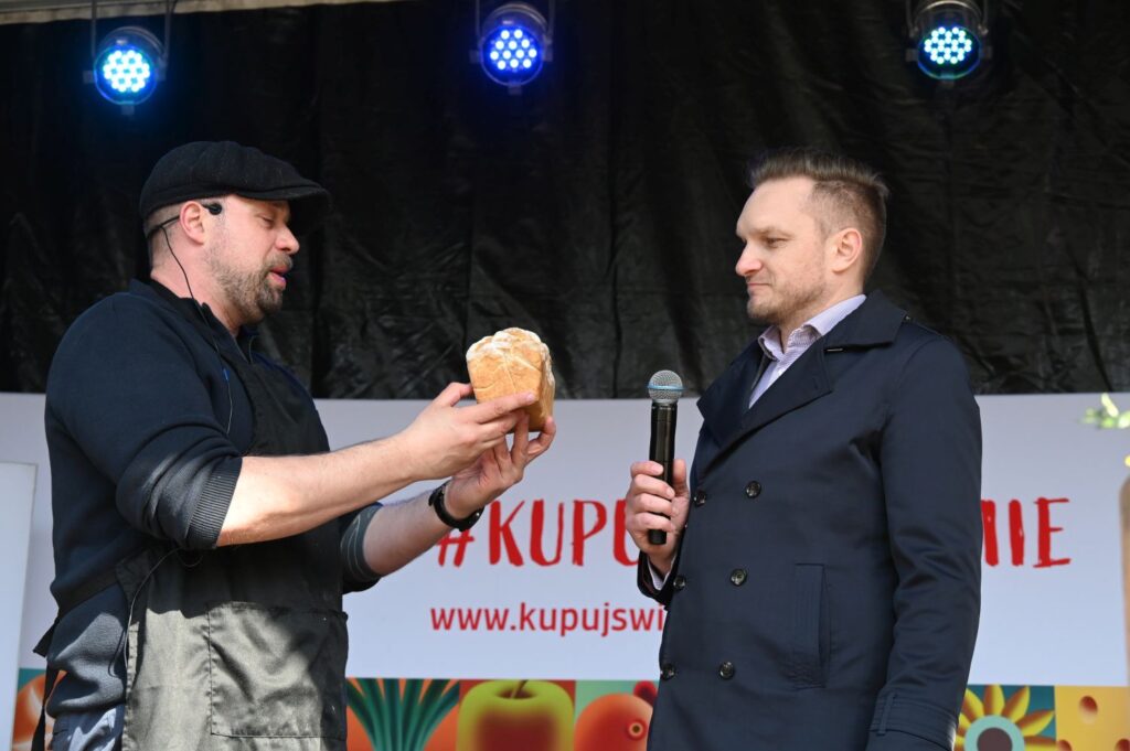 Mężczyzna na scenie prezentuje wypiek drugiemu uczestnikowi podczas konkursu kulinarnego (kliknięcie powiększy zdjęcie).
