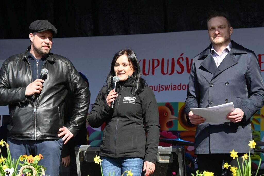 Troje prowadzących stoi na scenie z mikrofonami podczas wręczenia nagród w konkursie na wypiek (kliknięcie powiększy zdjęcie).