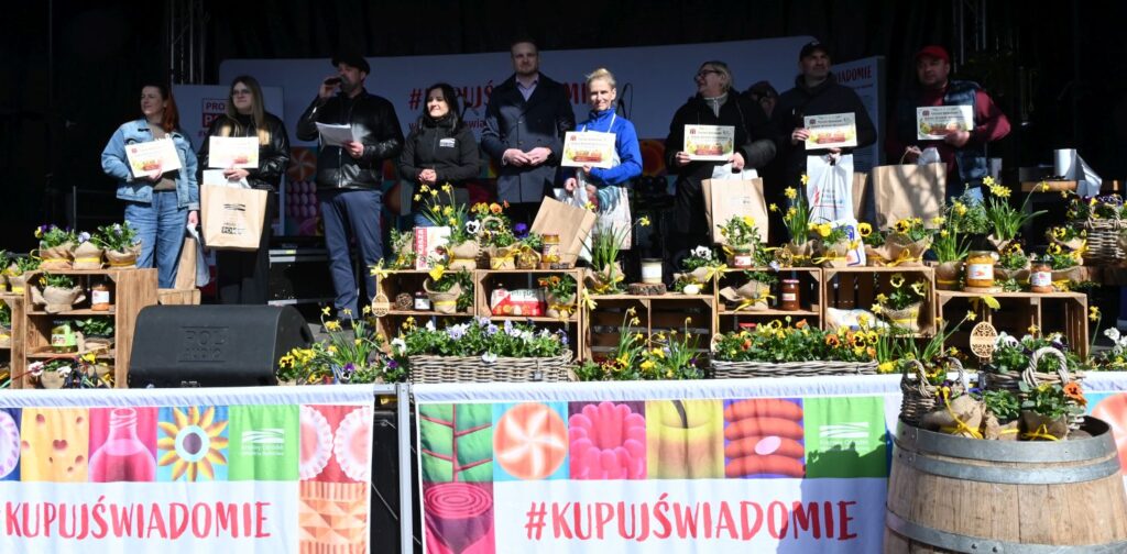 Grupa laureatów konkursu na wypiek stoi na scenie z dyplomami i nagrodami, na tle dekoracji wiosennych i stoiska z produktami (kliknięcie powiększy zdjęcie).