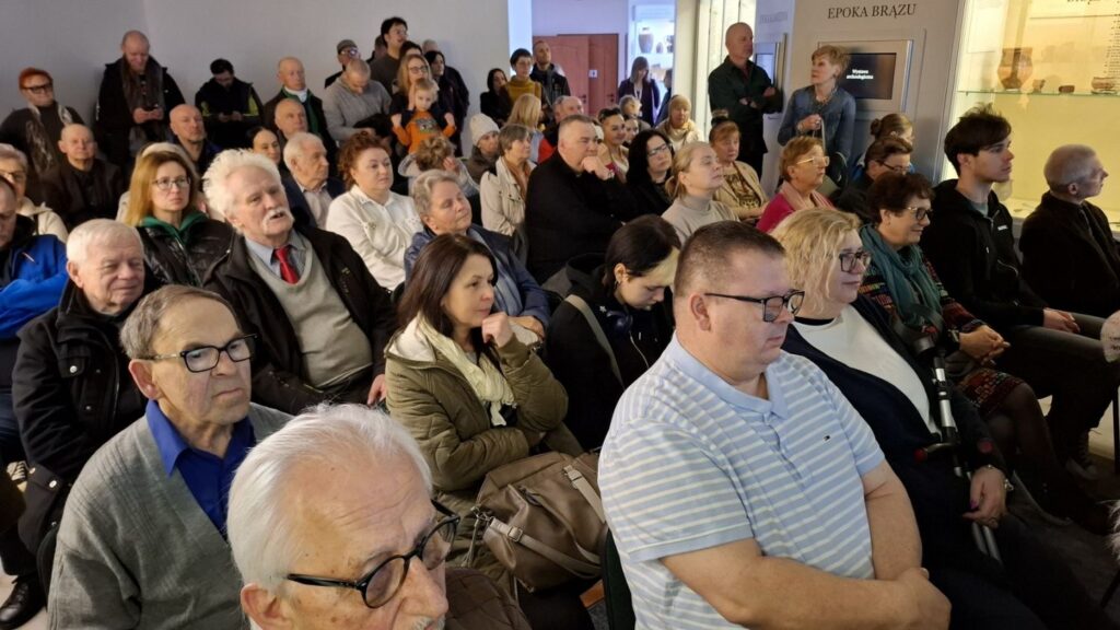 Uczestnicy wydarzenia siedzą blisko siebie, skupieni na słuchaniu prezentacji (kliknięcie powiększy zdjęcie).