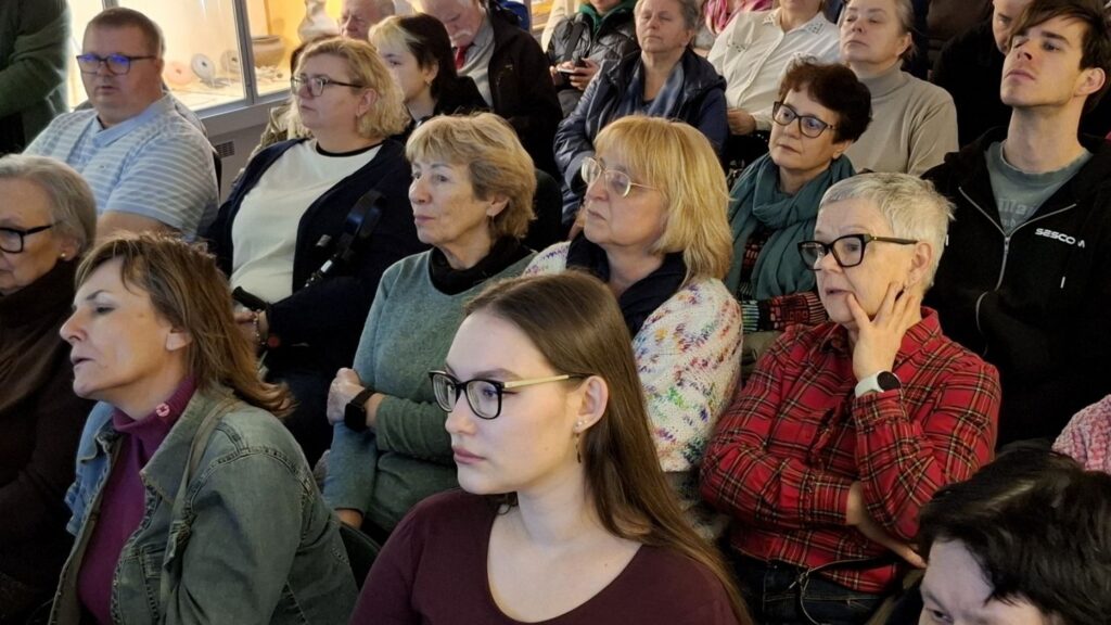 Zbliżenie na słuchaczy wykładu, wśród nich osoby w różnym wieku uważnie słuchające prelegenta (kliknięcie powiększy zdjęcie).