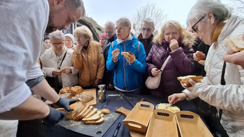 Zbliżenie na degustację chleba – prowadzący kroi pieczywo, a zgromadzeni uczestnicy próbują jego kawałków (kliknięcie powiększy zdjęcie).