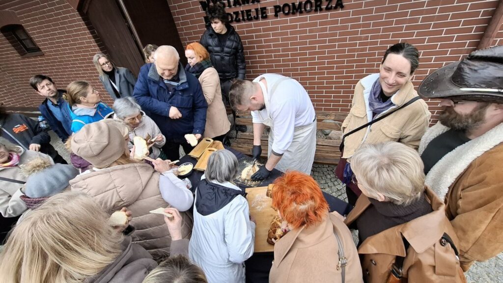 Uczestnicy wydarzenia stoją wokół stołu przed budynkiem i obserwują przygotowywanie oraz krojenie chleba (kliknięcie powiększy zdjęcie).
