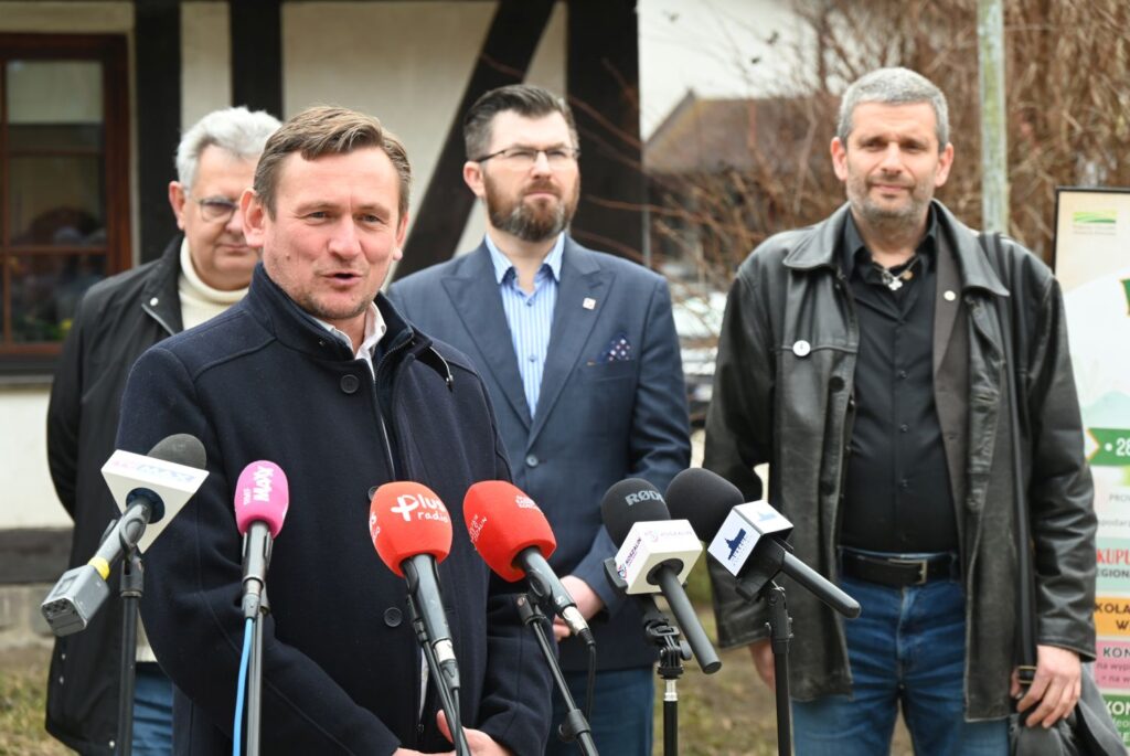 Prezydent Koszalina stoi przed mikrofonami i przemawia w trakcie konferencji prasowej, za nim stoi trzech uczestników konferencji (kliknięcie powiększy zdjęcie).