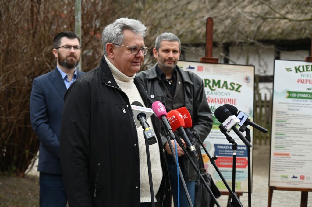 Dyrektor Bałtyckiego Teatru Dramatycznego przemawia do mikrofonów, w tle stojące na sztalugach plakaty wydarzenia i zabudowania zabytkowej chaty (kliknięcie powiększy zdjęcie).