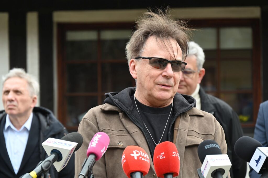 Mężczyzna w przyciemnionych okularach udzielają wypowiedzi mediom podczas konferencji prasowej, w tle widoczni inni uczestnicy wydarzenia (kliknięcie powiększy zdjęcie).