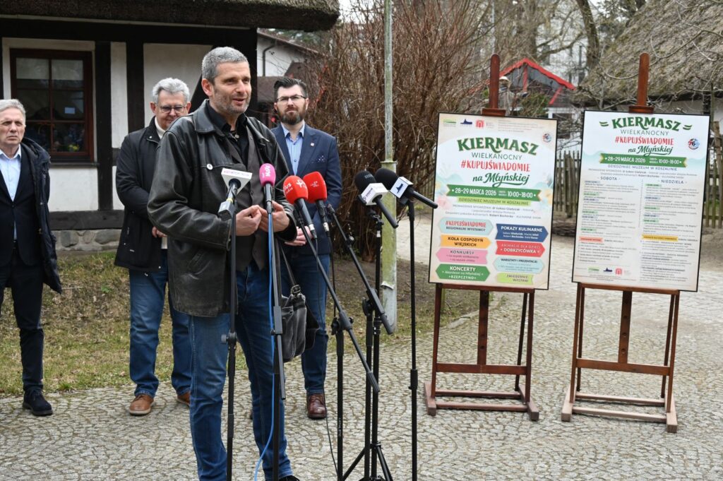 Mężczyzna przemawiający przy mikrofonach na tle plansz informacyjnych dotyczących wydarzenia, obok stoją uczestnicy konferencji (kliknięcie powiększy zdjęcie).