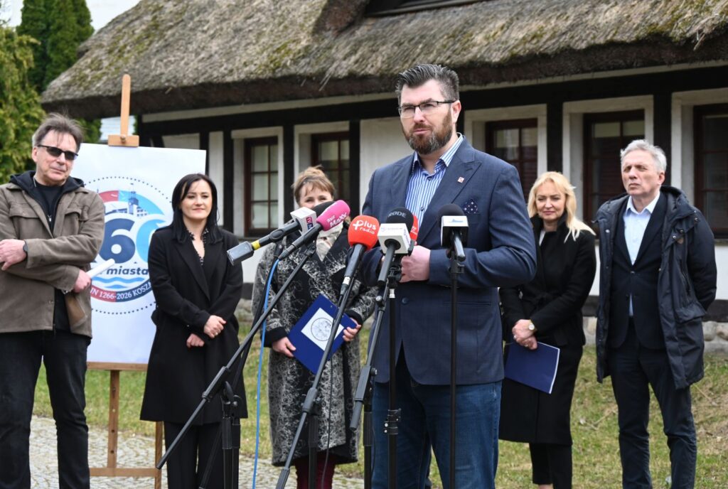 Mężczyzna przemawiający do mediów podczas konferencji prasowej na świeżym powietrzu, w tle widoczni organizatorzy i zabudowania biurowe muzeum (kliknięcie powiększy zdjęcie).