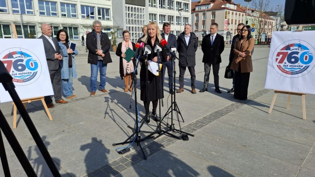 Kobieta przemawia do mikrofonów a za nią stoją samorządowcy i przedstawiciele instytucji oraz plansza z logo 760-lecia miasta Koszalin (kliknięcie powiększy zdjęcie).