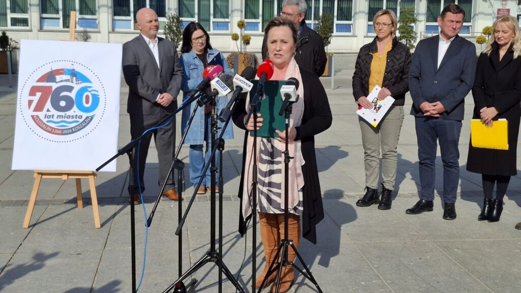 Dyrektor muzeum przemawia podczas konferencji prasowej na tle planszy z logo 760-lecia miasta Koszalin, za nią stoją przedstawiciele władz i współorganizatorzy wydarzenia (kliknięcie powiększy zdjęcie).