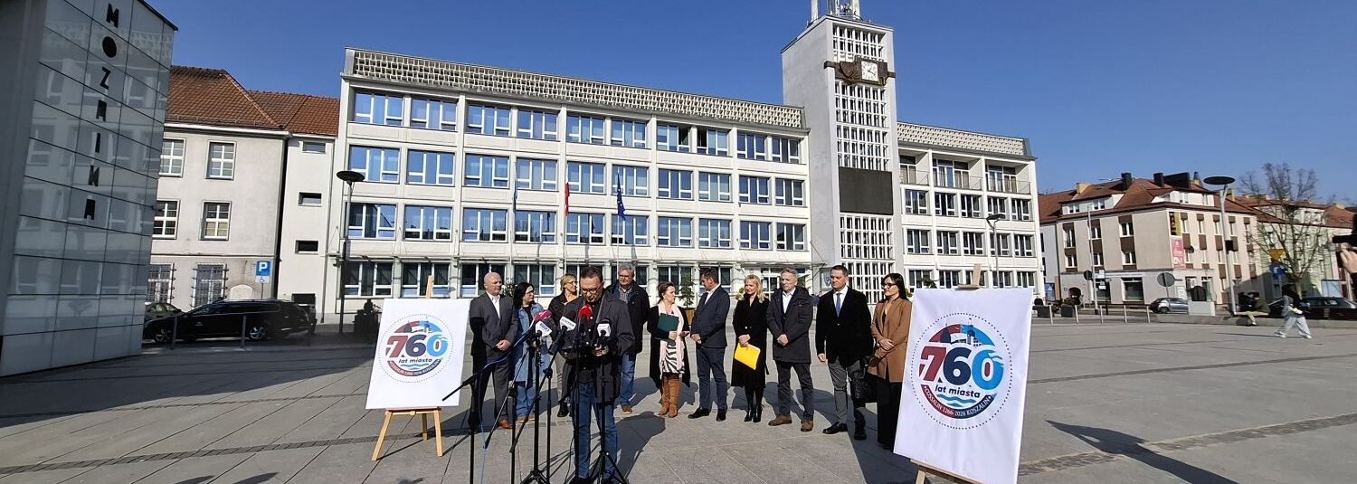 Szerokie ujęcie konferencji prasowej na placu przed ratuszem z widocznym budynkiem urzędu miasta Koszalin i grupą uczestników stojących przy planszach z logo 760-lecia miasta (kliknięcie powiększy zdjęcie).