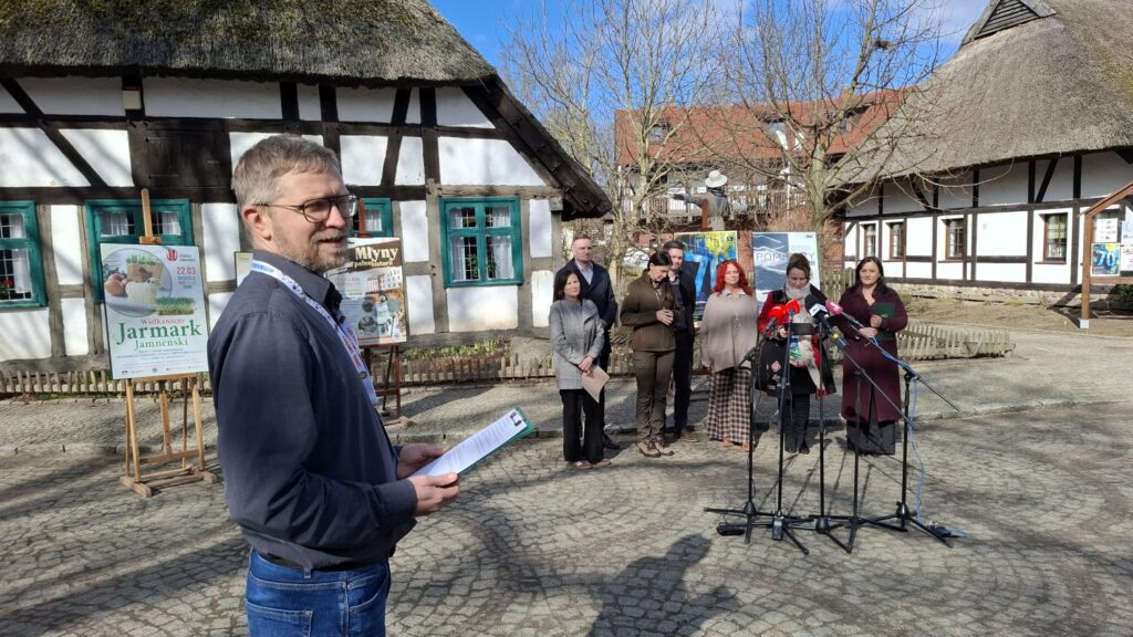 Mężczyzna prowadzący konferencję stoi z boku, przed zabytkową chałupą krytą strzechą, przed mikrofonami stoją przedstawiciele organizatorów wydarzeń (kliknięcie powiększy zdjęcie).