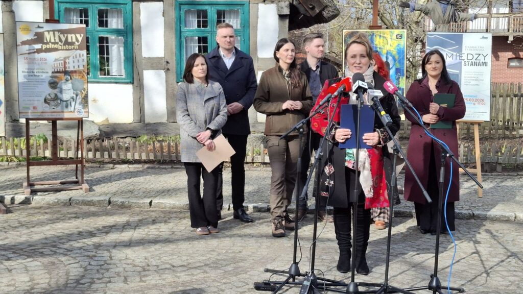 Joanna Chojecka przemawia do mikrofonów podczas konferencji prasowej przed budynkiem zabytkowej chałupy, a za nią stoją pozostali uczestnicy konferencji (kliknięcie powiększy zdjęcie).