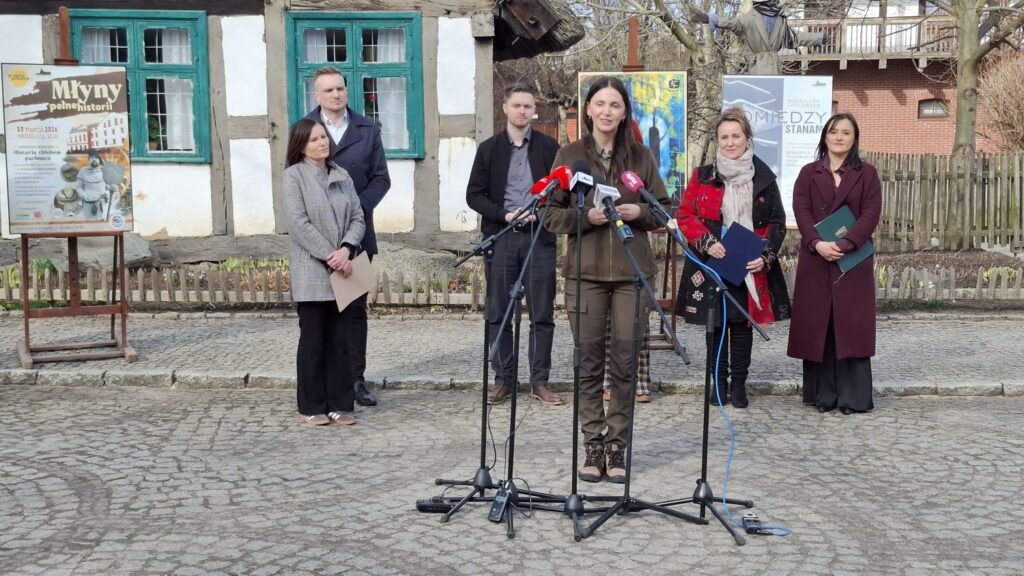 Dagny Nowak-Staszewska przemawia do mikrofonów podczas konferencji prasowej przed budynkiem zabytkowej chałupy, a za nią stoją pozostali uczestnicy konferencji (kliknięcie powiększy zdjęcie).
