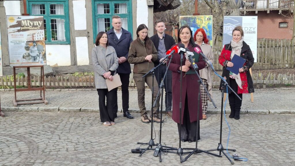 Dorota Stawiszyńska przemawia do mikrofonów podczas konferencji prasowej przed budynkiem zabytkowej chałupy, a za nią stoją pozostali uczestnicy konferencji (kliknięcie powiększy zdjęcie).