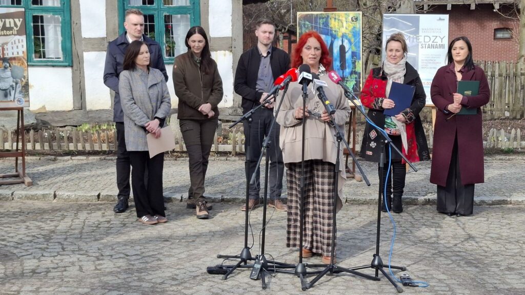 Ewa Miśkiewicz-Żebrowska przemawia do mikrofonów podczas konferencji prasowej przed budynkiem zabytkowej chałupy, a za nią stoją pozostali uczestnicy konferencji (kliknięcie powiększy zdjęcie).