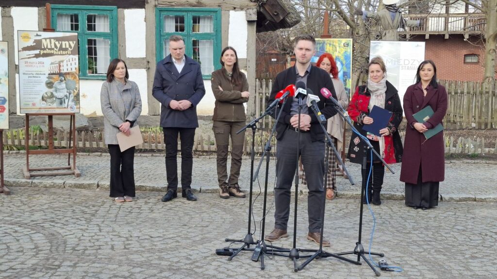 Łukasz Rozmarynowski przemawia do mikrofonów podczas konferencji prasowej przed budynkiem zabytkowej chałupy, a za nim stoją pozostali uczestnicy konferencji (kliknięcie powiększy zdjęcie)