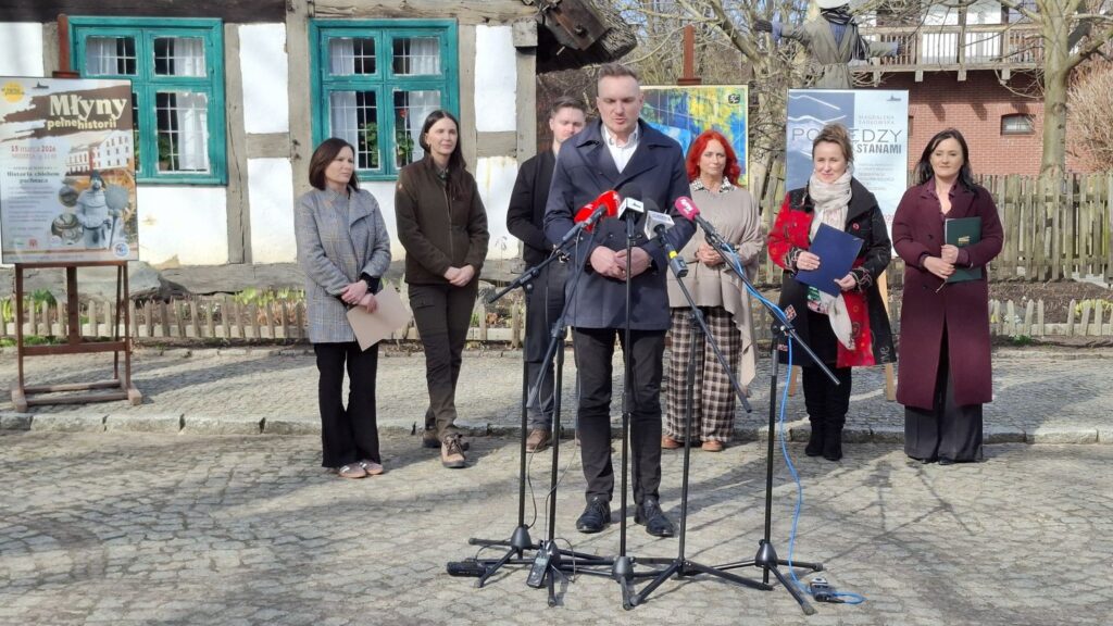 Piotr Kyczka przemawia do mikrofonów podczas konferencji prasowej przed budynkiem zabytkowej chałupy, a za nim stoją pozostali uczestnicy konferencji (kliknięcie powiększy zdjęcie).