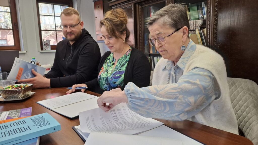 Trzy osoby siedzą przy stole i przeglądają dokumenty oraz książki historyczne, w tle umeblowanie biura (kliknięcie powiększy zdjęcie).