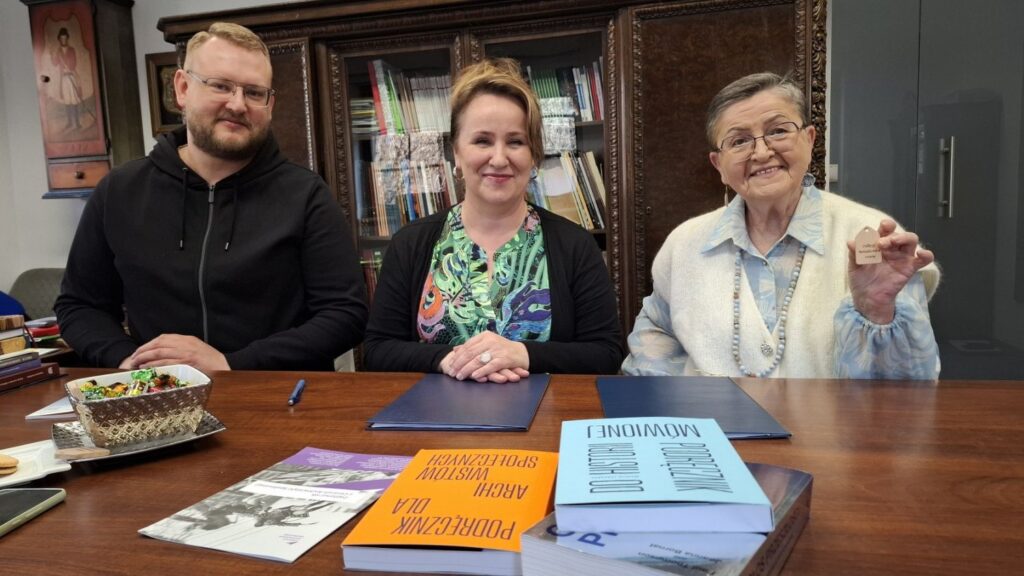 Trzy osoby siedzą przy stole z książkami i dokumentami, patrzą w stronę aparatu, a jedna z kobiet trzyma w dłoni niewielki przedmiot (kliknięcie powiększy zdjęcie).