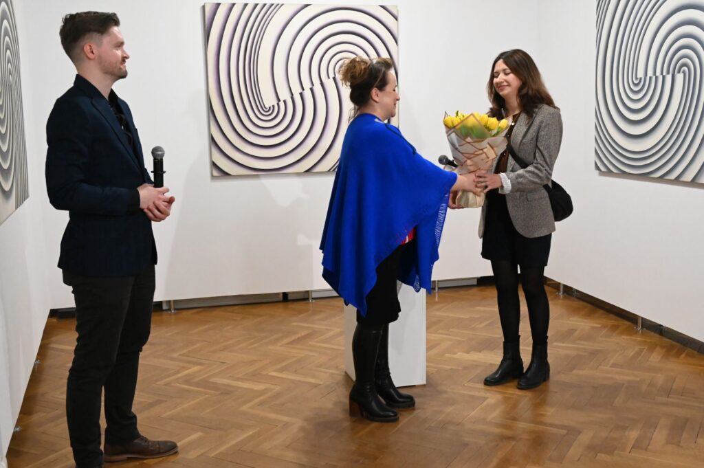 Dyrektor muzeum wręcza kobiecie bukiet kwiatów podczas wernisażu, w tle widoczne abstrakcyjne obrazy na ścianie galerii (kliknięcie powiększy zdjęcie).