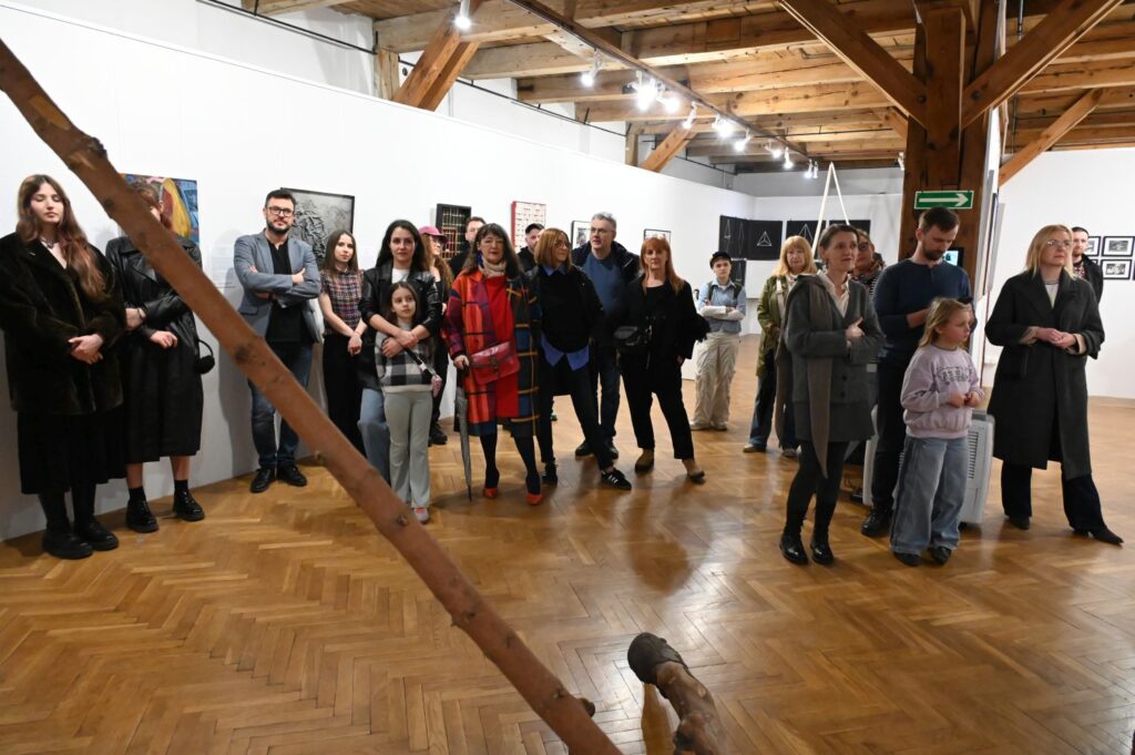 Grupa gości zgromadzona w sali wystawowej podczas wernisażu, widoczne drewniane belki i przestrzeń sali wystawowej (kliknięcie powiększy zdjęcie).