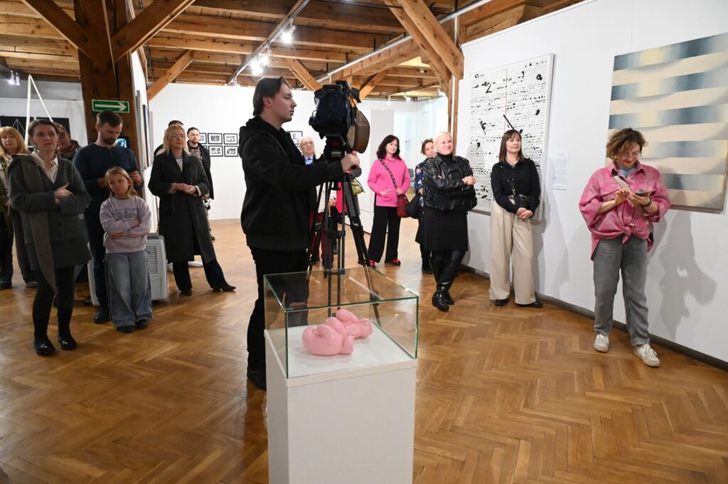 Osoba filmująca wydarzenie kamerą oraz uczestnicy stojący na sali wystawowej, na pierwszym planie przeszklona gablota z artefaktem wystawy (kliknięcie powiększy zdjęcie).