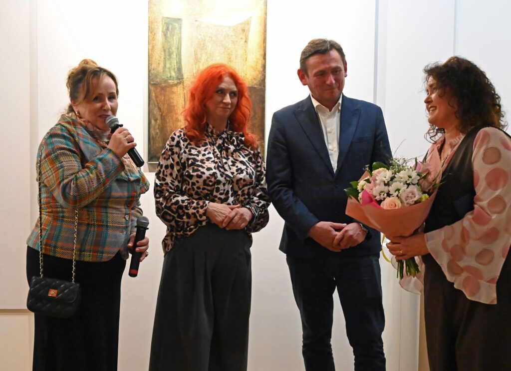 Przemówienie podczas wernisażu – kobieta z mikrofonem w towarzystwie trzech osób przemawia do zaproszonych gości (kliknięcie powiększy zdjęcie).