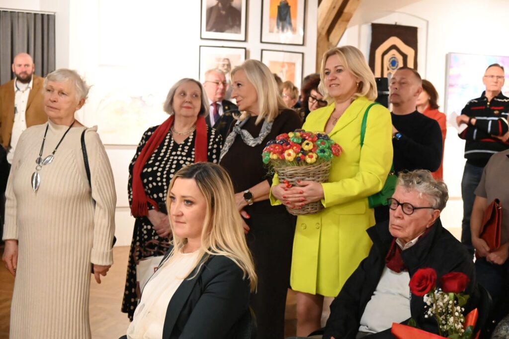 Uczestnicy wydarzenia słuchający przemówień podczas wernisażu wystawy, w środku stoi kobieta trzymająca w rękach bukiet kwiatów (kliknięcie powiększy zdjęcie).