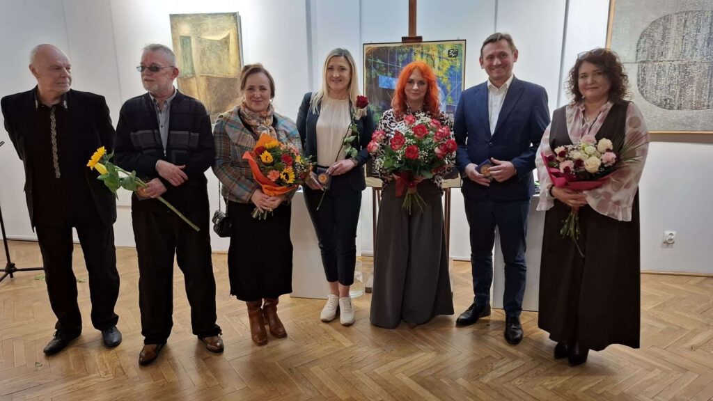 Siedem osób pozuje do wspólnego zdjęcia w sali wystawowej na tle obrazów, kilka z nich trzyma w rękach bukiety kwiatów (kliknięcie powiększy zdjęcie).
