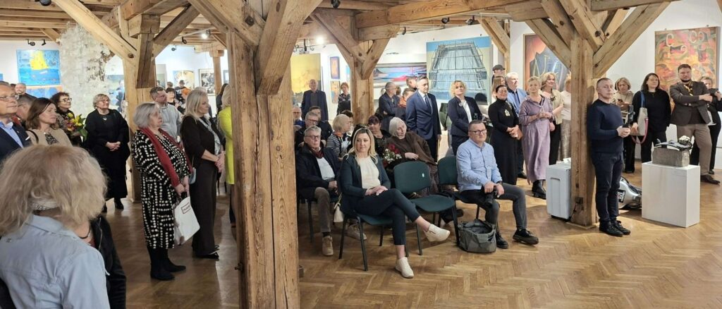 Licznie zgromadzona publiczność siedzi i stoi w przestrzeni sali podczas wernisażu wystawy (kliknięcie powiększy zdjęcie).