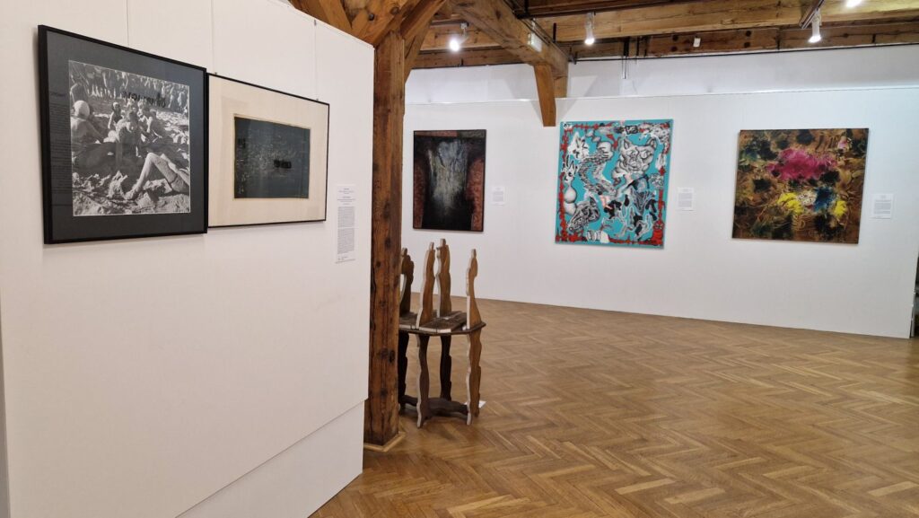 Sala wystawowa z prezentacją współczesnych prac – na białych ścianach wiszą obrazy i fotografie, a na środku stoi niewielki stolik z rzeźbiarskimi formami, widoczna drewniana konstrukcja stropu galerii (kliknięcie powiększy zdjęcie).