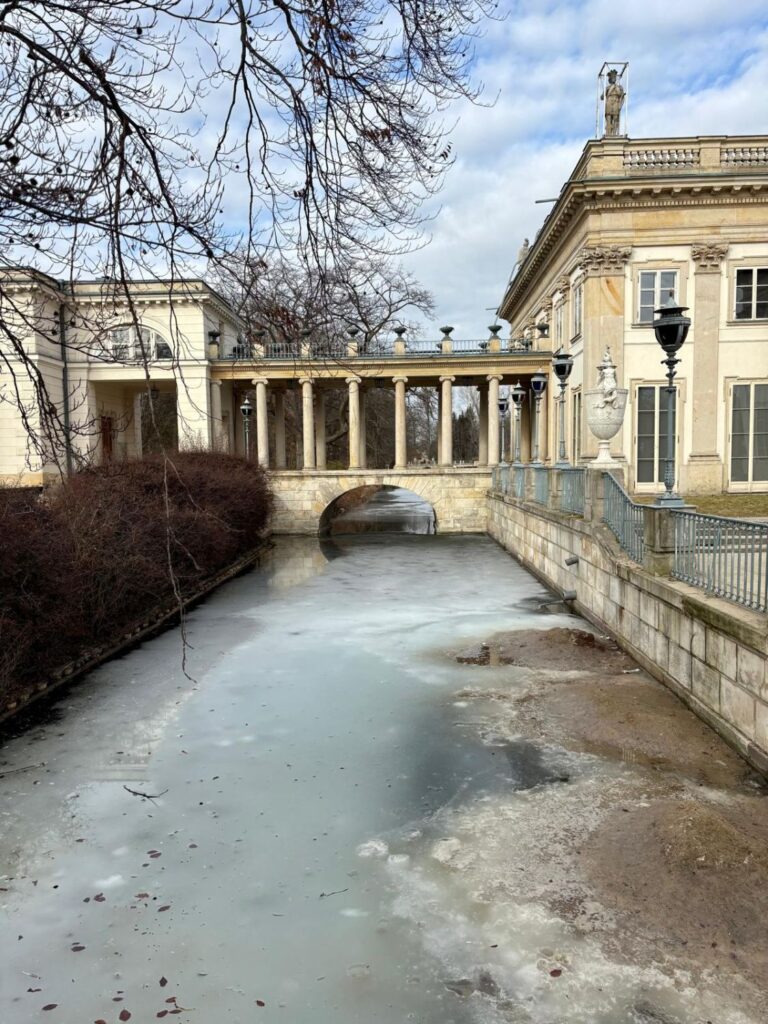 Klasycystyczna kolumnada nad kanałem w parku, pod którą przepływa częściowo zamarznięta woda, a po bokach widać ozdobne balustrady i fragment pałacowej zabudowy (kliknięcie powiększy zdjęcie).