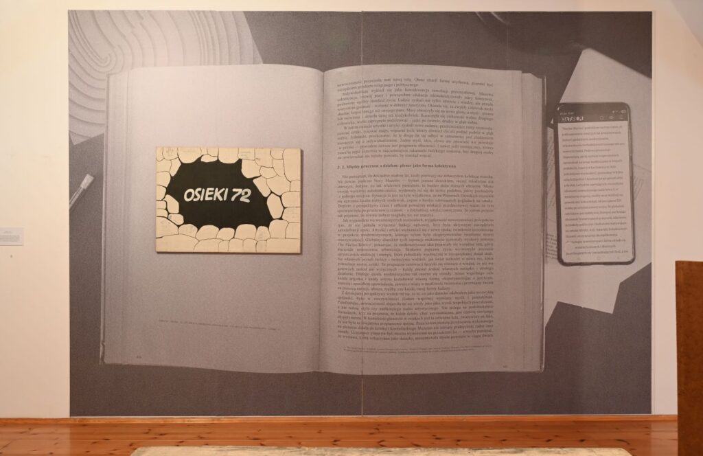 Duża ścienna grafika przedstawiająca otwartą książkę z napisem „Osieki 72” eksponowana na białej ścianie galerii (kliknięcie powiększy zdjęcie).