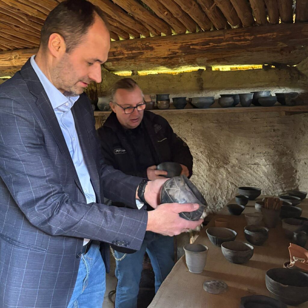 Dwóch mężczyzn we wnętrzu chaty ogląda i trzyma w rękach gliniane naczynia ustawione na stole i półkach (kliknięcie powiększy zdjęcie).