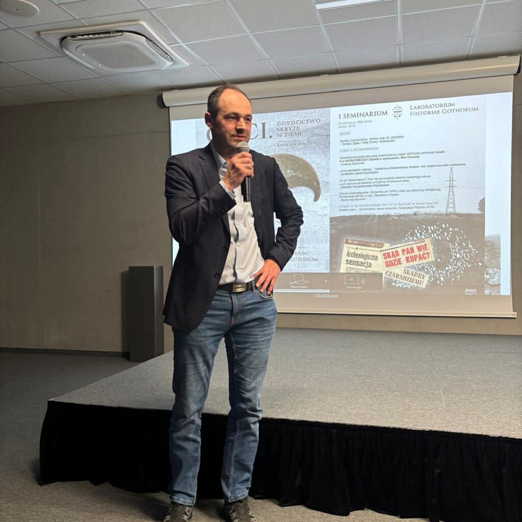 Mężczyzna stoi na scenie i przemawia do publiczności podczas konferencji, trzymając mikrofon, na tle ekranu z prezentacją (kliknięcie powiększy zdjęcie).