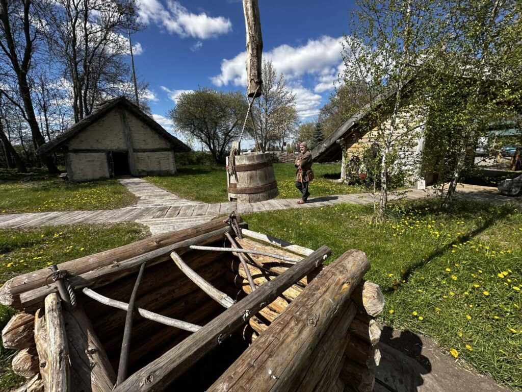 Drewniana studnia z żurawiem stoi na pierwszym planie, w tle widoczne są zabudowania skansenu i ścieżka wśród zieleni w słoneczny dzień (kliknięcie powiększy zdjęcie).