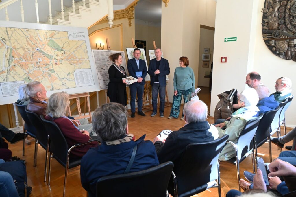 Przywitanie gości w sali muzealnej – prelegenci stoją przed dużą mapą i przemawiają do siedzącej publiczności (kliknięcie powiększy zdjęcie).