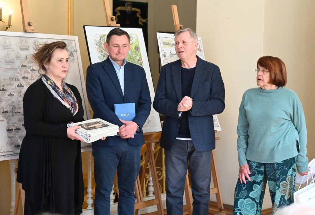 Kilka osób stoi przy planszach z ilustracjami, jedna z kobiet trzyma w rękach publikacje i prezentuje je uczestnikom (kliknięcie powiększy zdjęcie).