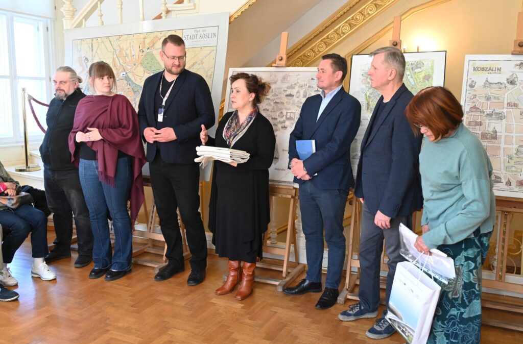 Grupa organizatorów i uczestników wydarzenia stoi w rzędzie przy planszach wystawowych w sali muzeum (kliknięcie powiększy zdjęcie).