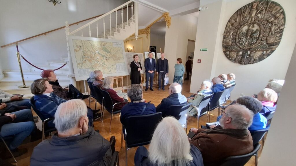 Uczestnicy wydarzenia siedzą na krzesłach i słuchają wystąpienia w przestronnej sali z widoczną dużą mapą na ścianie (kliknięcie powiększy zdjęcie).