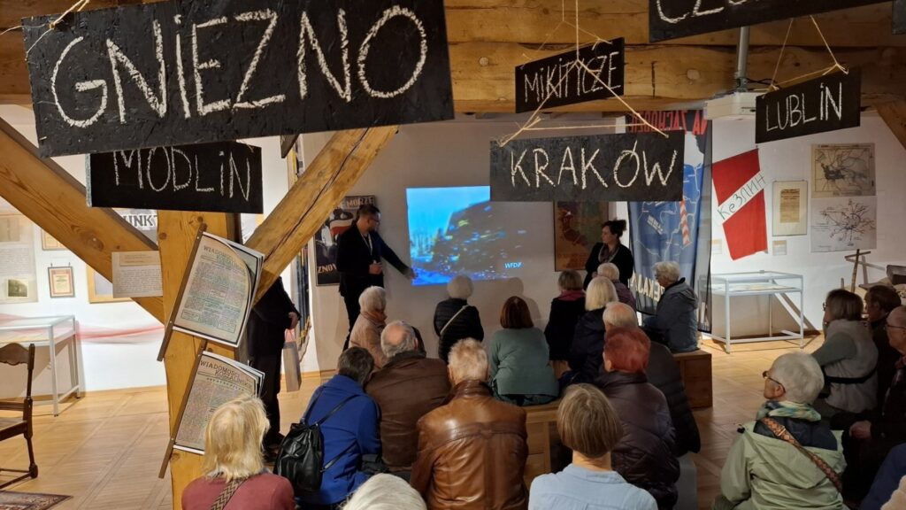 Wnętrze ekspozycji z drewnianą konstrukcją, uczestnicy siedzą i oglądają prezentację multimedialną (kliknięcie powiększy zdjęcie).