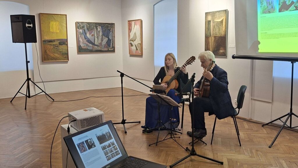 Dwoje muzyków siedzi na krzesłach i gra na gitarach podczas wydarzenia, tle obrazy na ścianach (kliknięcie powiększy zdjęcie).