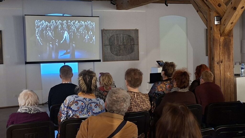 Publiczność ogląda projekcję filmu wyświetlanego na ekranie w sali galerii (kliknięcie powiększy zdjęcie).