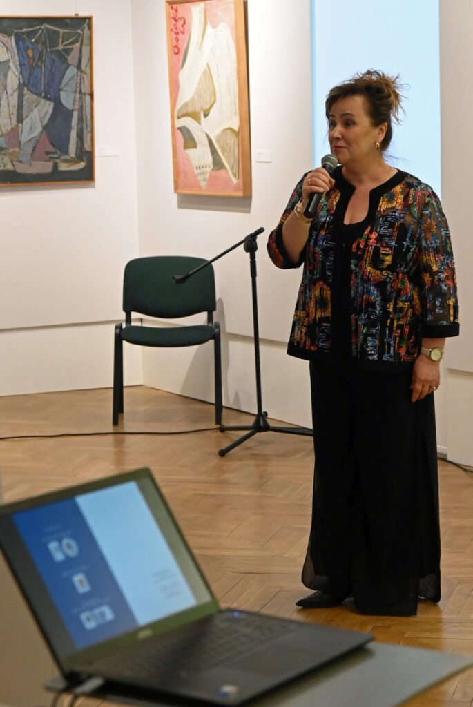 Kobieta przemawia do publiczności, trzymając mikrofon, w tle widoczne obrazy na ścianach galerii (kliknięcie powiększy zdjęcie).