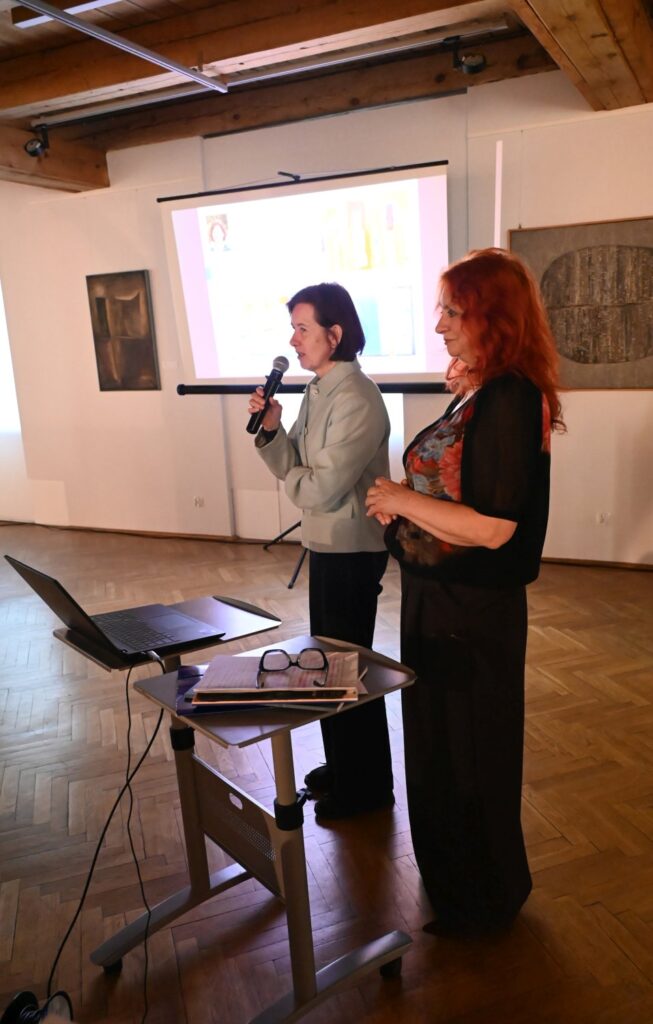 Dwie kobiety stoją przy stoliku z laptopem, jedna przemawia do publiczności z mikrofonem, w tle ekran projekcyjny (kliknięcie powiększy zdjęcie).
