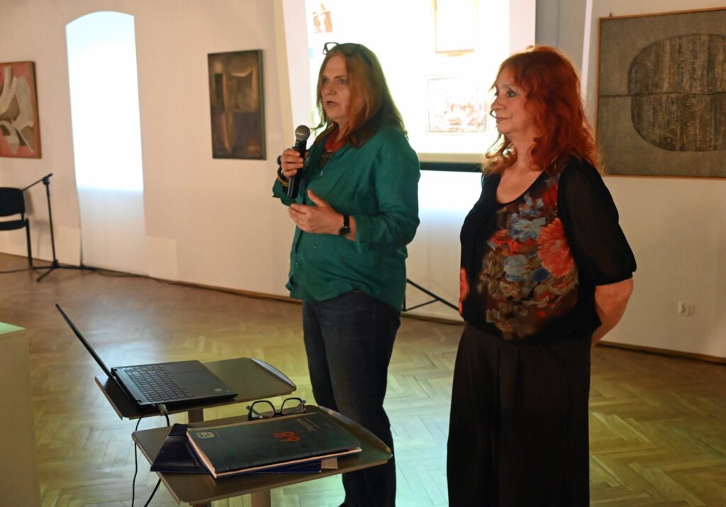 Dwie kobiety stoją obok siebie, jedna przemawia do zgromadzonych, w tle widoczne obrazy na ścianach galerii (kliknięcie powiększy zdjęcie).