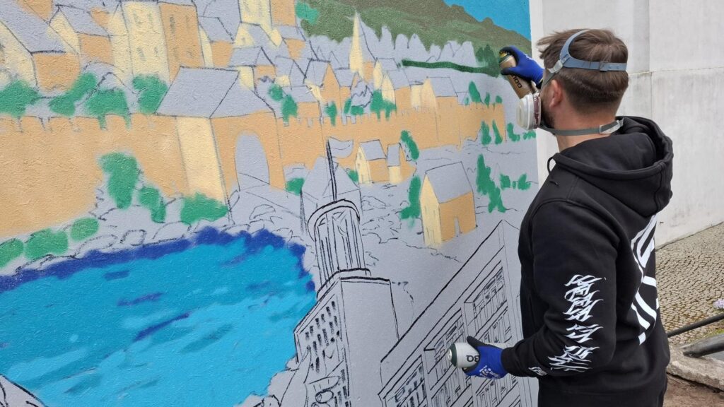 Mężczyzna w czarnej bluzie tworzy graffiti na dużej planszy z widokiem miasta. Widoczne są szkice budynków oraz elementy malowane sprayem. (kliknięcie powiększy zdjęcie).