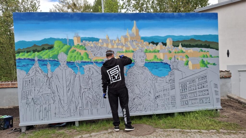 Artysta stoi przed niemal ukończonym muralem przedstawiającym miejską panoramę i historyczne postacie podczas pracy nad detalami graffiti. (kliknięcie powiększy zdjęcie).